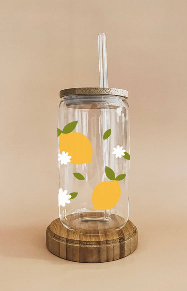 A clear glass cup with a bamboo lid and straw is decorated with yellow lemon slices, green leaves, and white daisy decals, resting on a round wooden base.
